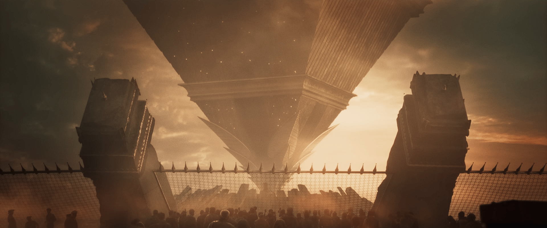 A massive triangular megastructure rises above a crowd of people gathered near a fortified gate at sunset. Named COMPLEX
