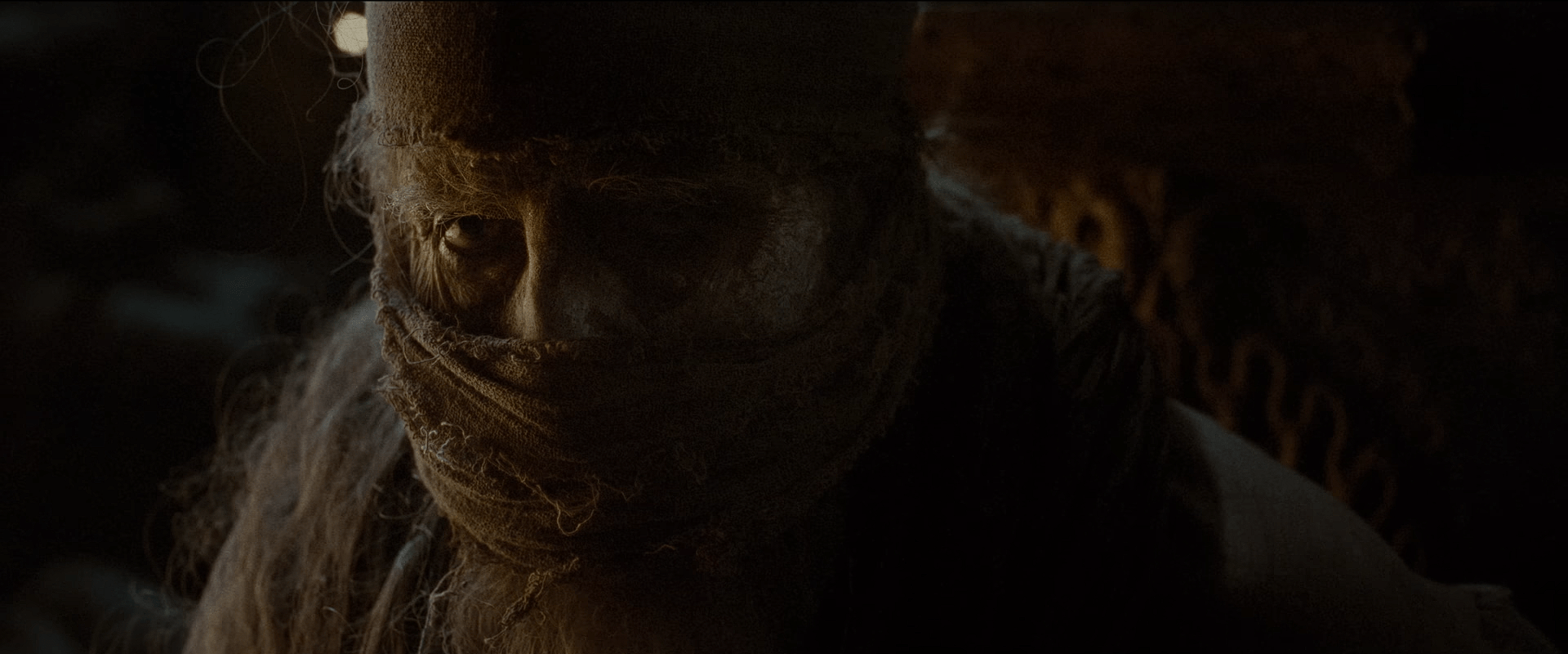 A mysterious hooded figure with cloth wrapped over the lower face watches intently from the shadows.