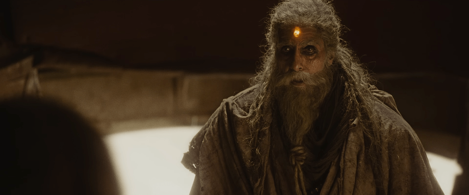 An elderly, bearded figure with glowing light on the forehead stands in a dim chamber, wearing worn, ancient-looking robes.