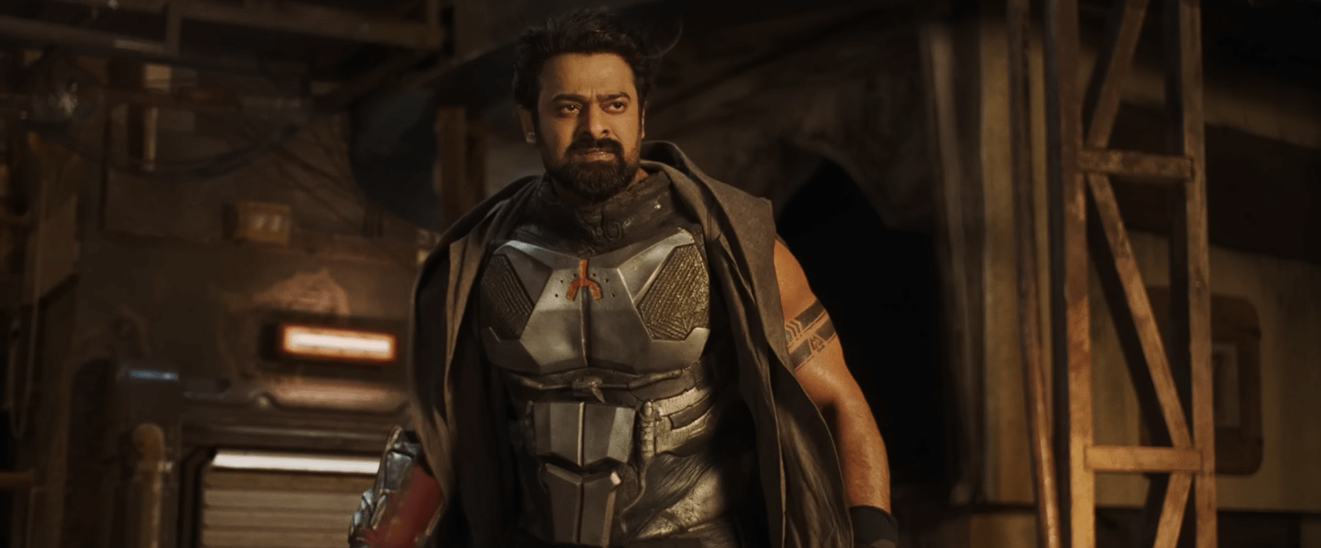 A rugged man in metallic armor and a cloak stands confidently in a dusty, industrial settlement.