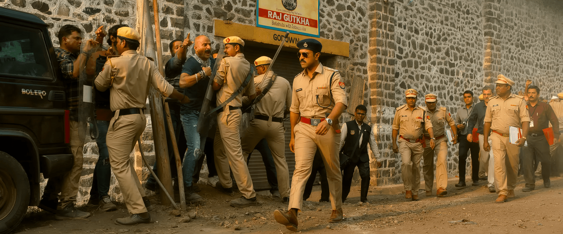 Ram Charan, appearing as a police officer IPS, leads a team of uniformed officers during a raid as they detain criminals near a warehouse-like building.