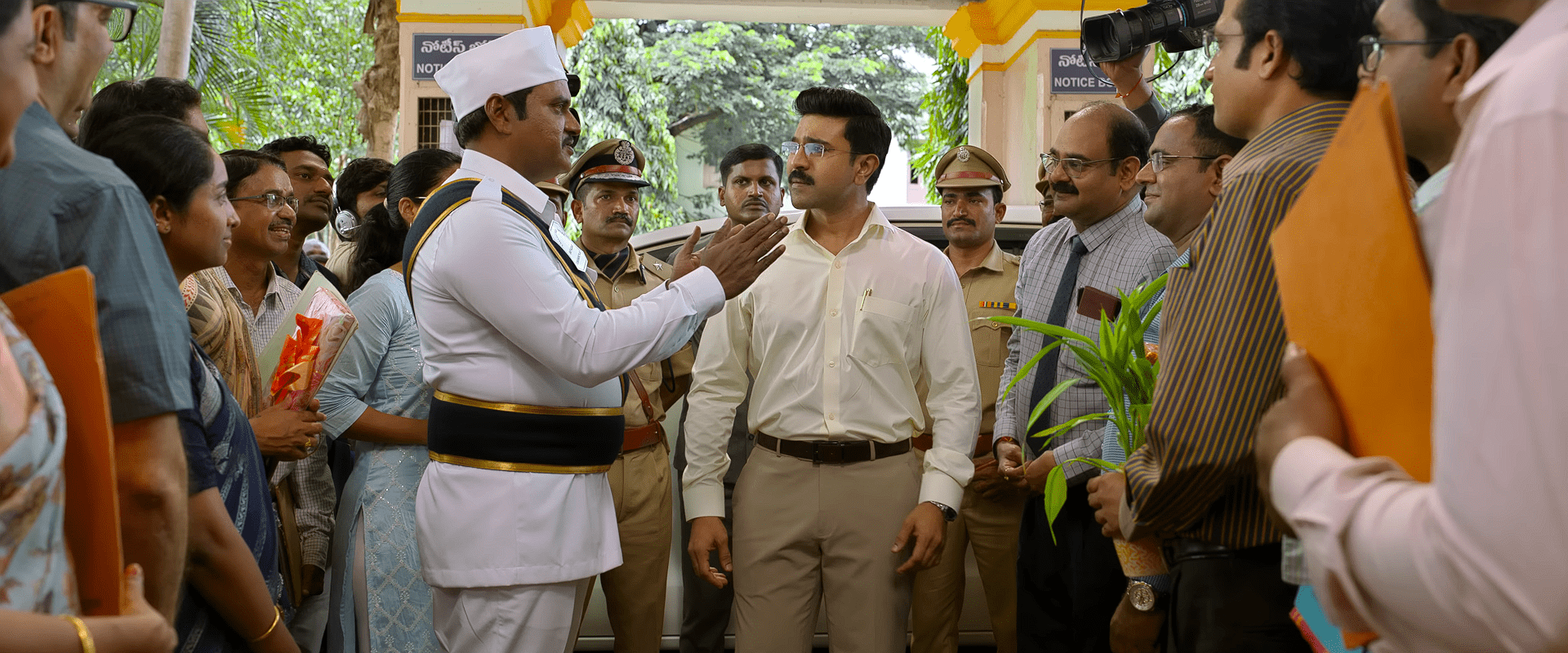 Ram Charan, in an IAS-style formal outfit, listens as a uniformed officer speaks to him amid a crowd of officials, media, and onlookers outside a government office.