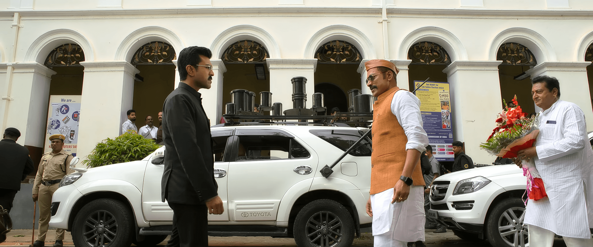 Ram Charan, dressed in black formal attire, faces S. J. Suryah, who arrives wearing a white kurta and orange vest outside a government building, surrounded by security and staff.