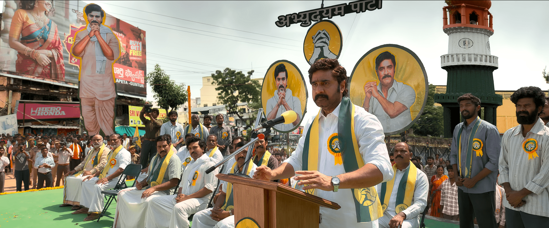 Srikanth stands at a political rally stage giving a speech, with large cut-outs of his character displayed behind him, while supporters and party members sit and stand around.
