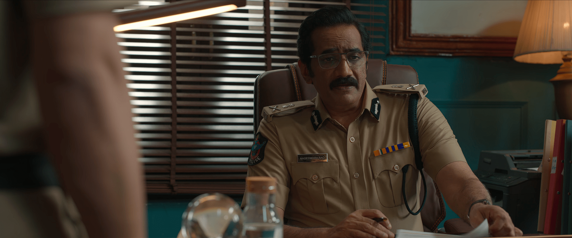 An older police officer, played by Rao Ramesh, sits behind a desk in uniform, looking serious while reviewing documents.