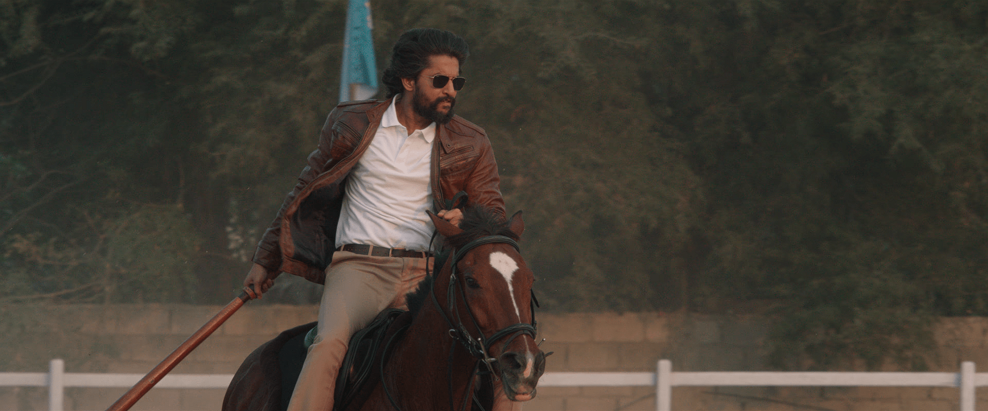 Nani rides a galloping horse while holding a wooden weapon, wearing sunglasses, a brown leather jacket, and beige trousers.