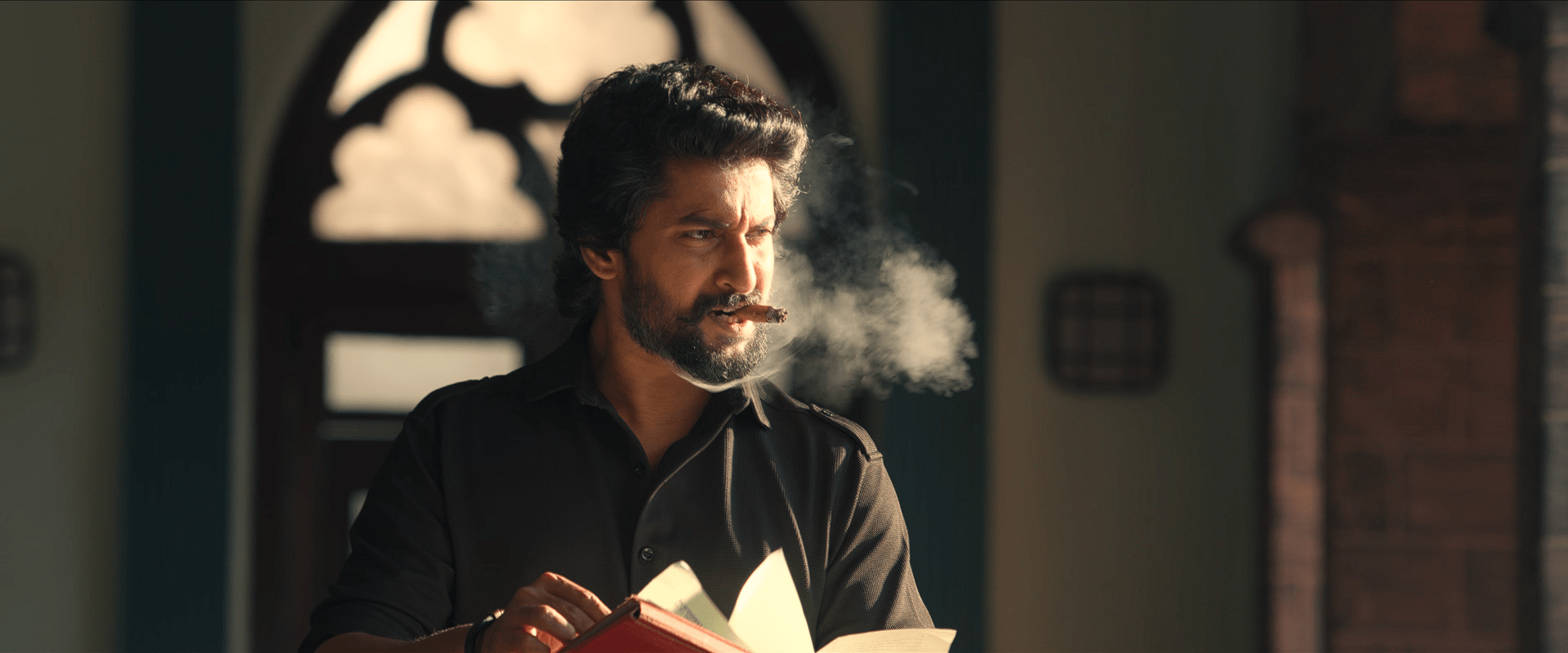 Nani smokes a cigar while flipping through a red book, sunlight streaming through an arched window behind him.