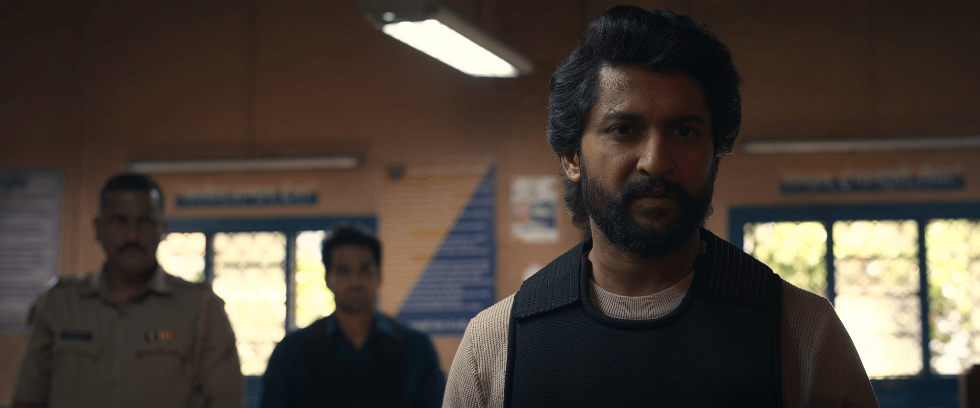 Nani, wearing a protective vest, looks serious inside a police station as two officers stand blurred behind him.