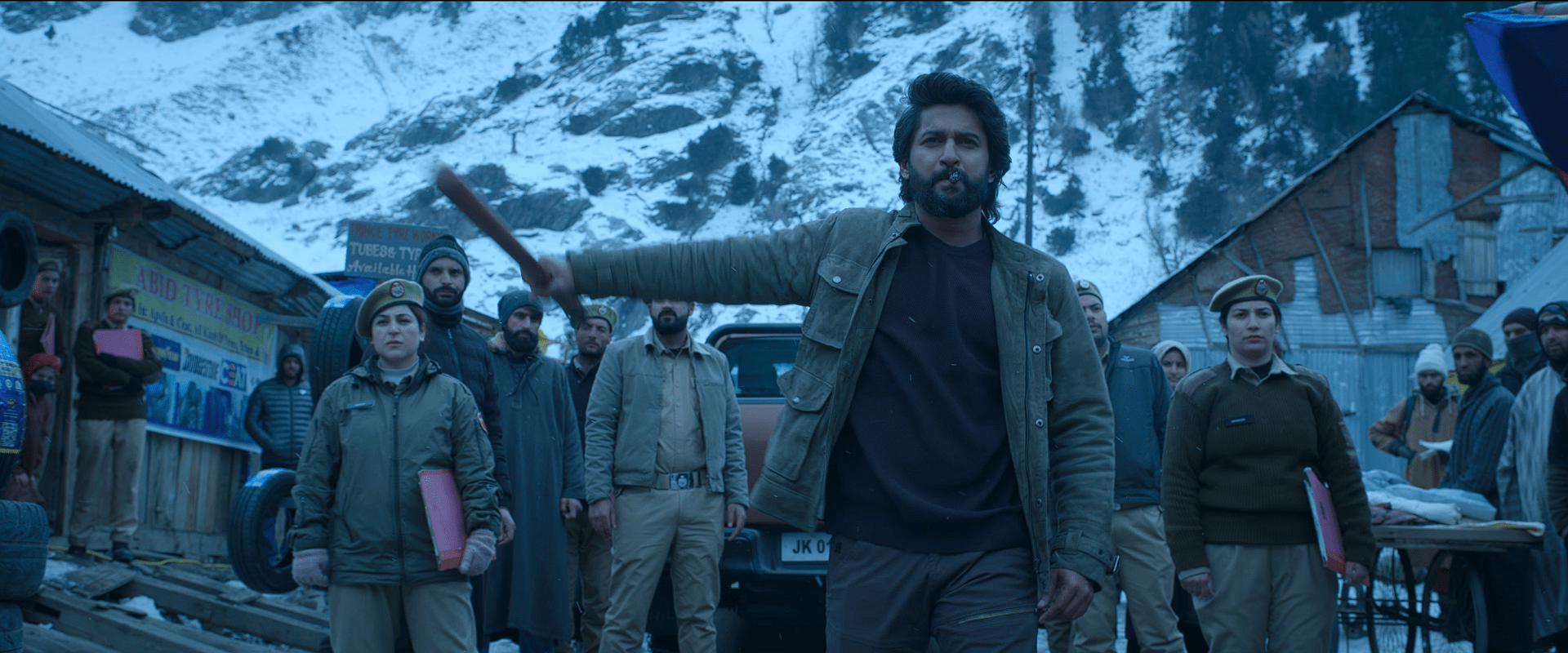 Nani stands confidently with a cigar in his mouth, holding a wooden stick while surrounded by police officers in a snowy mountain village backdrop.