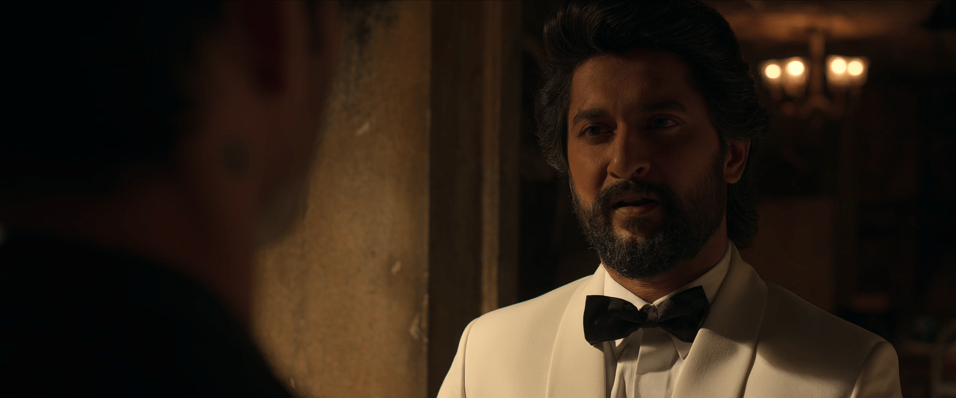 Nani, dressed in a white tuxedo with a black bow tie, speaks intensely to a man standing in front of him in a dim, warm-lit room.