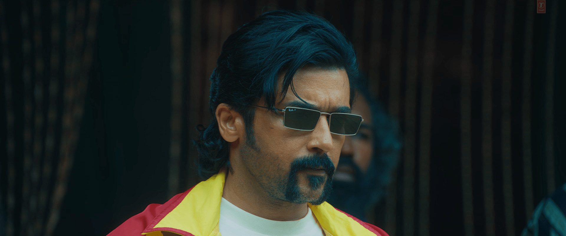 A stylish close-up of Suriya wearing narrow rectangular sunglasses and a red-yellow jacket, looking intensely ahead with a serious expression.