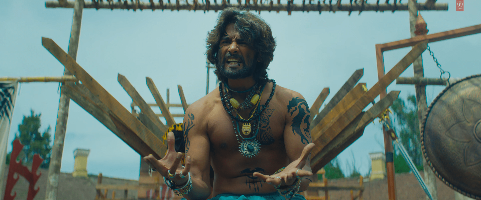 A dramatic shot of a muscular, tattooed tribal-warrior character shouting emotionally, adorned with beaded necklaces and traditional accessories, sitting against a backdrop of wooden weapons and tribal decor.