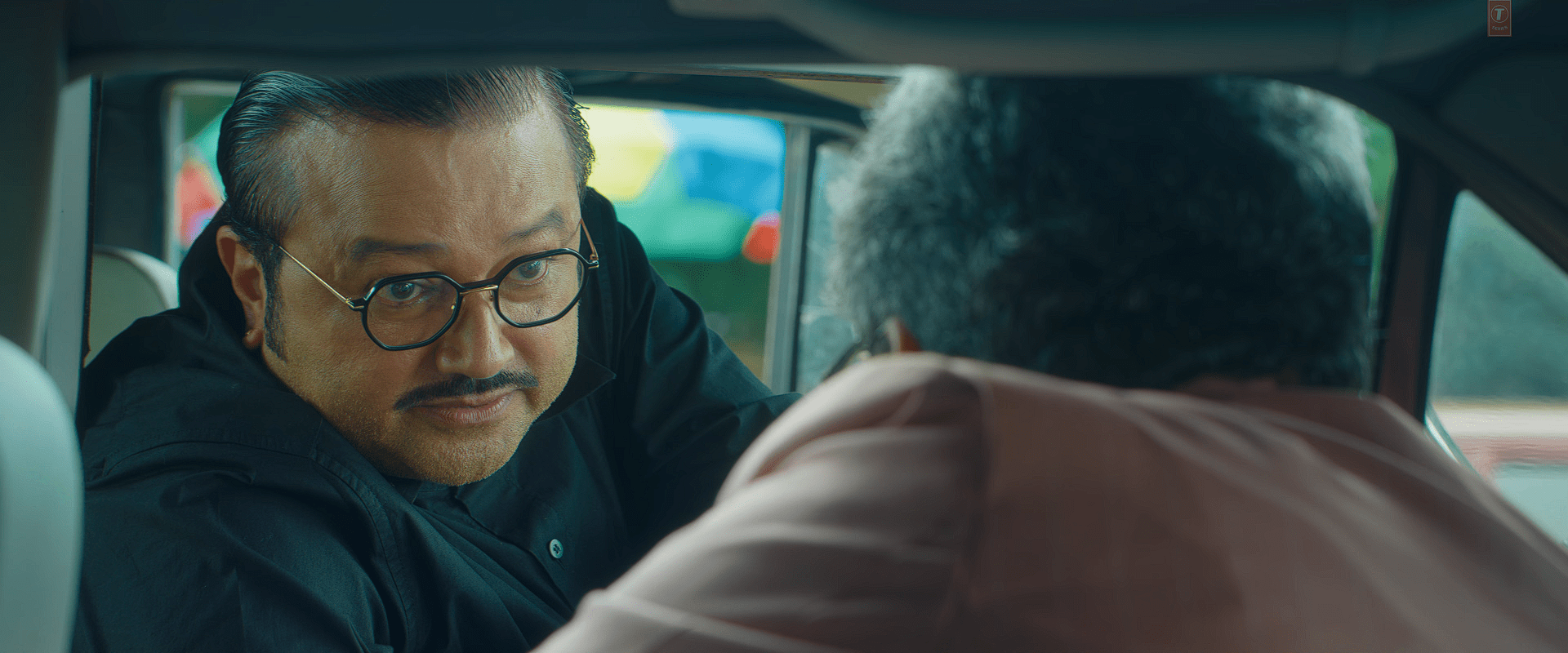 A medium close-up of a male character, leaning forward inside a car window with a stern expression, wearing glasses and a black outfit during an intense conversation.