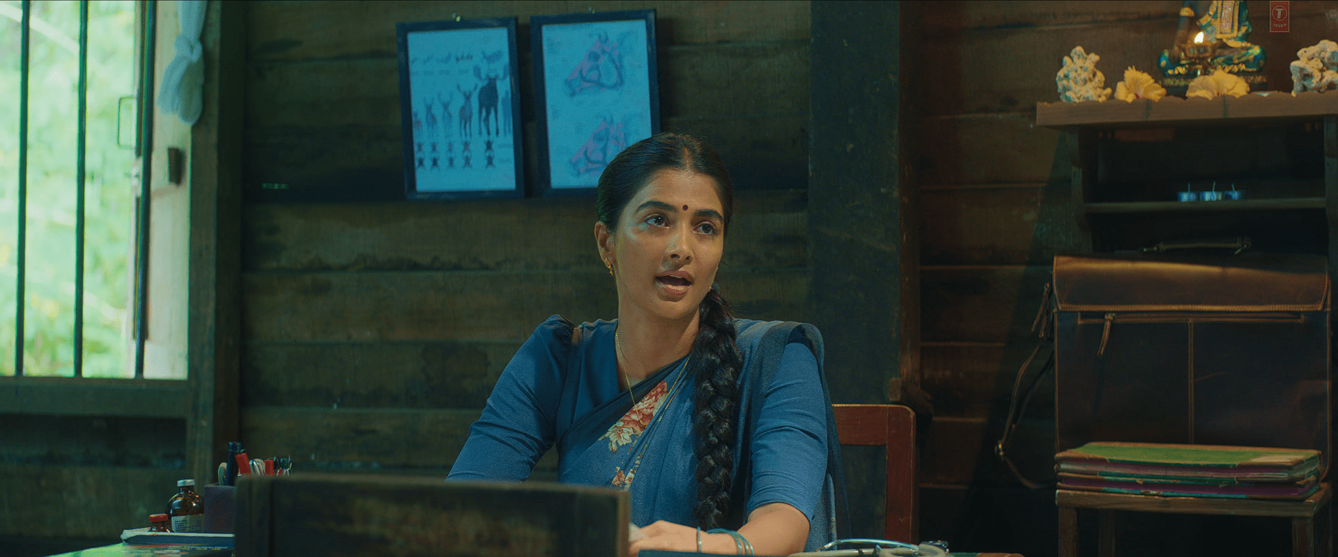 A close-up of Pooja Hegde sitting at a wooden desk in a dim, rustic office, wearing a blue saree with a long braid, speaking with a serious expression as daylight filters through the window beside her.