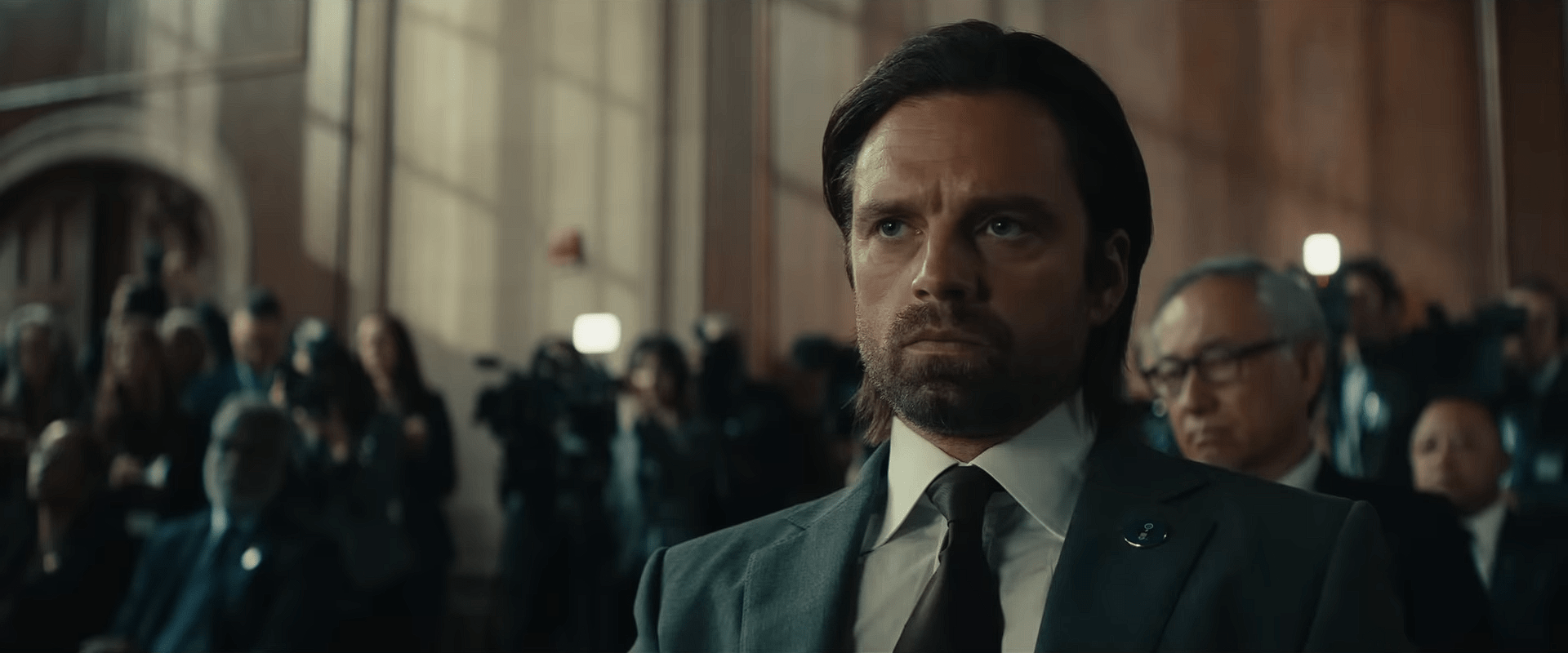 Sebastian Stan as Bucky Barnes sitting in a formal hearing room, wearing a suit and staring intensely while journalists and officials sit behind him.