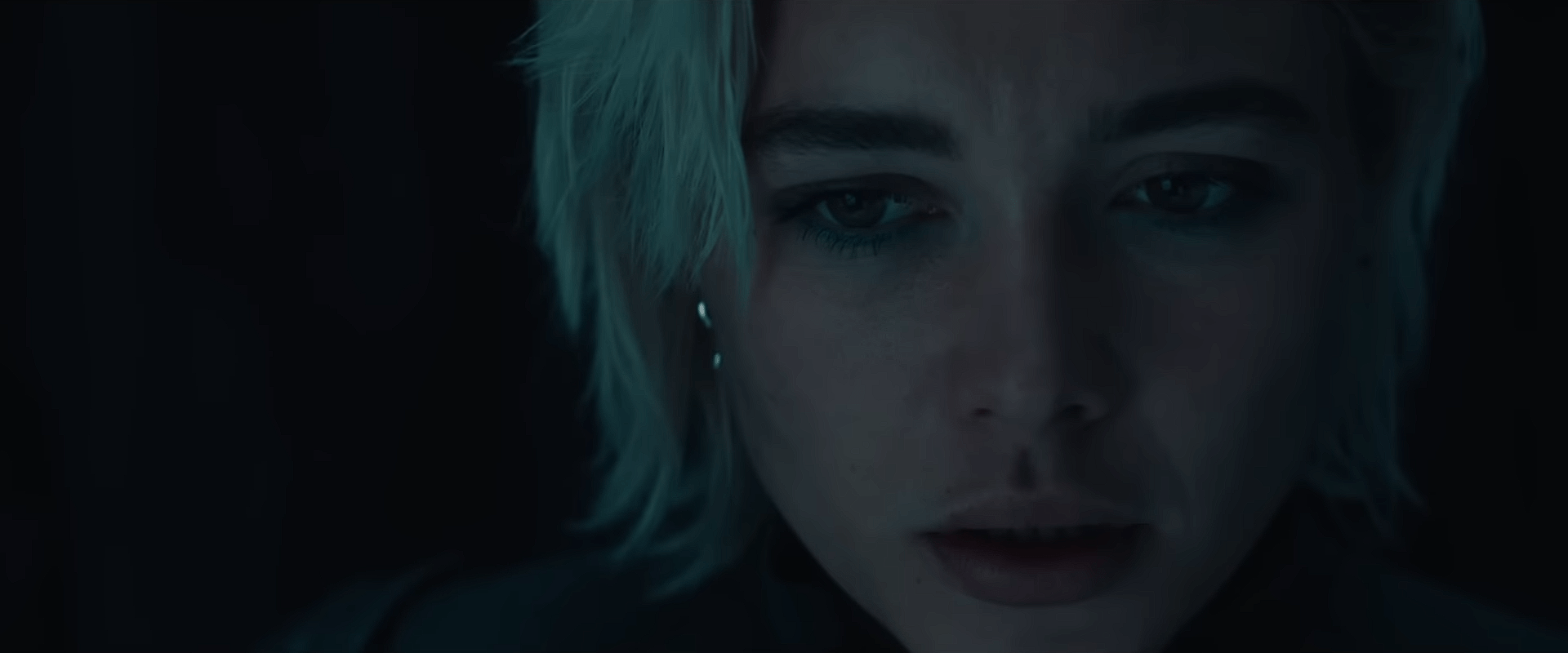 A close-up of Florence Pugh as Yelena Belova, her face lit dimly as she looks downward with a tense, emotional expression.