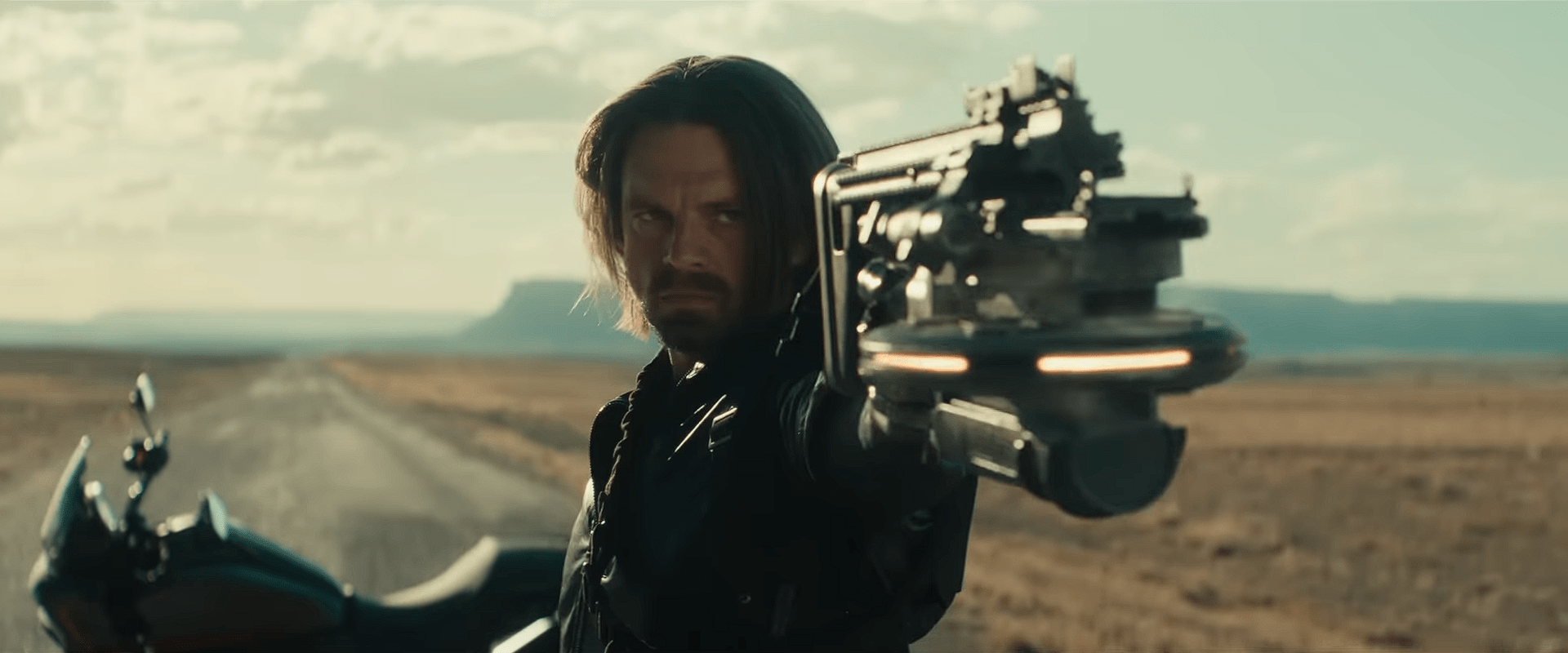 A close-up shot of Bucky Barnes, played by Sebastian Stan, standing on a deserted road and aiming a high-tech weapon forward, with his motorcycle parked behind him.