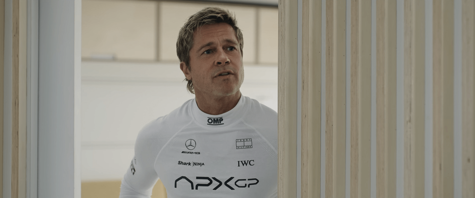F1 driver in a white racing undersuit standing near a wooden partition, appearing focused and preparing before the race.