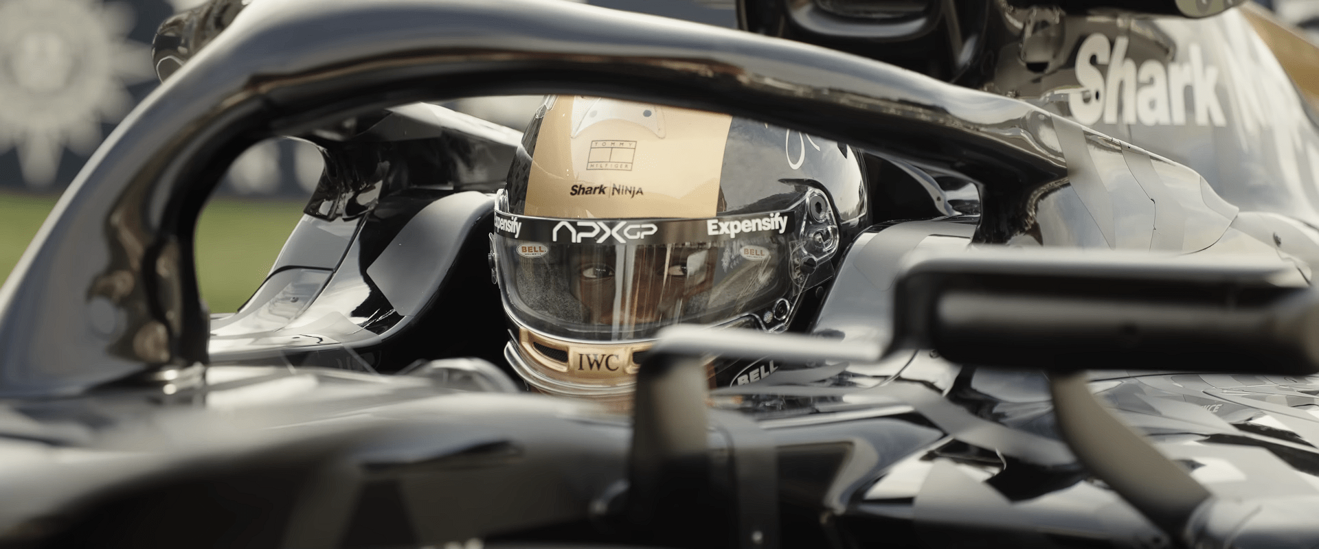 Close-up view of an F1 driver wearing a gold racing helmet, seen through the halo safety bar while seated inside the car cockpit.