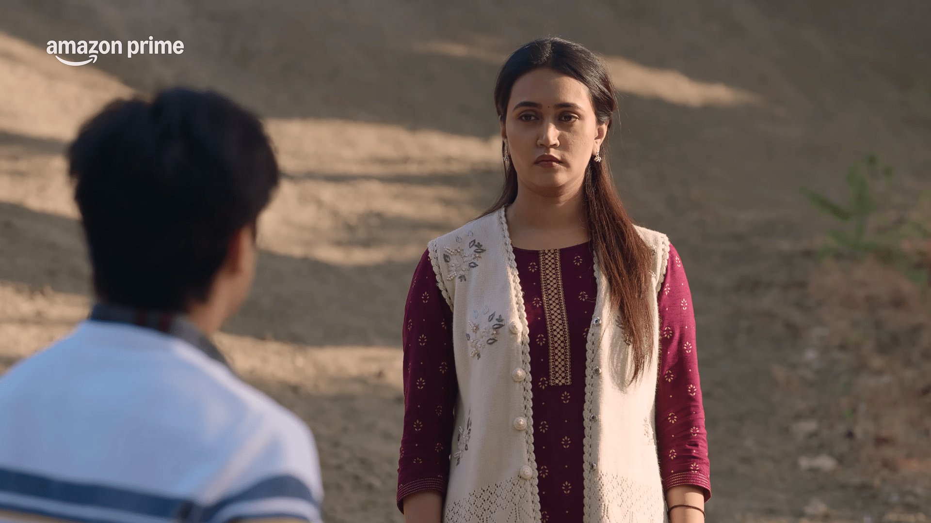 A young woman in a maroon kurta and embroidered white vest stands facing a man whose back is visible, her expression calm but serious, outdoors on a sunny day.