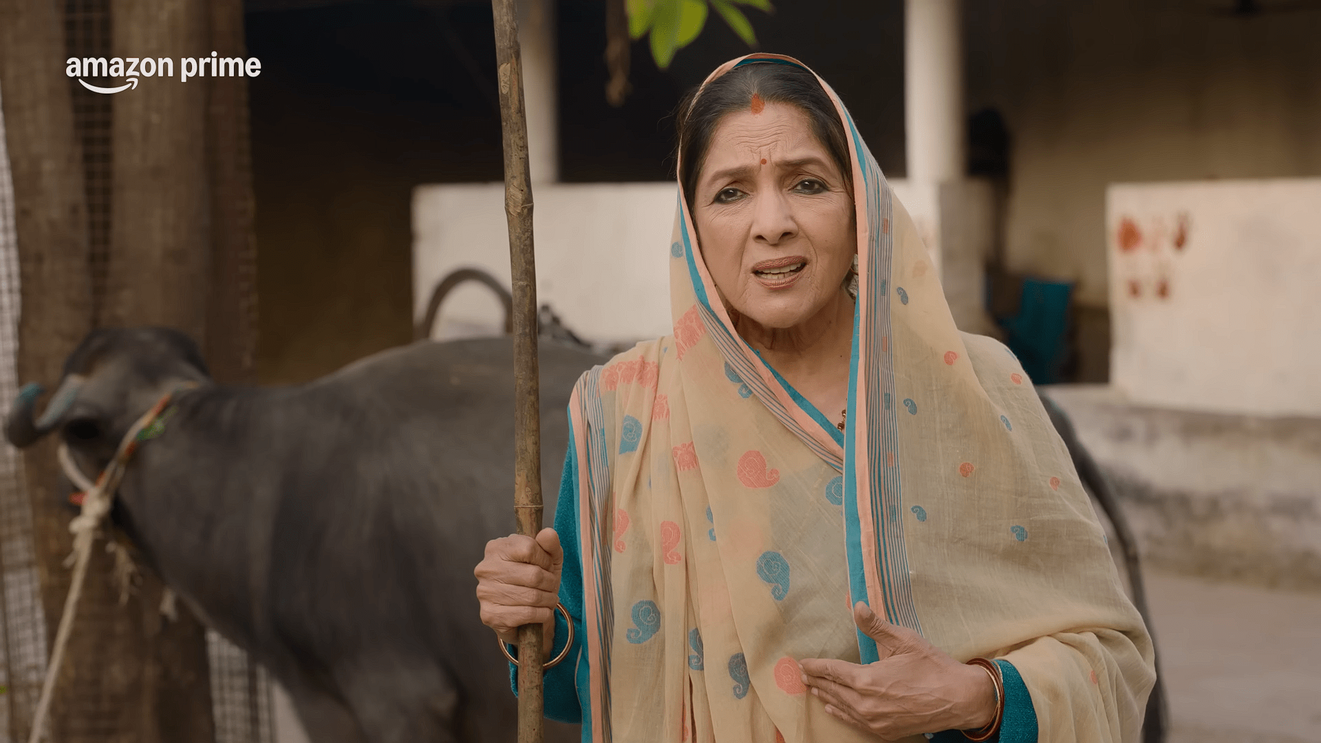Pradhan's Wife holding a wooden stick speaks with concern, standing beside a tied buffalo in a rural courtyard.