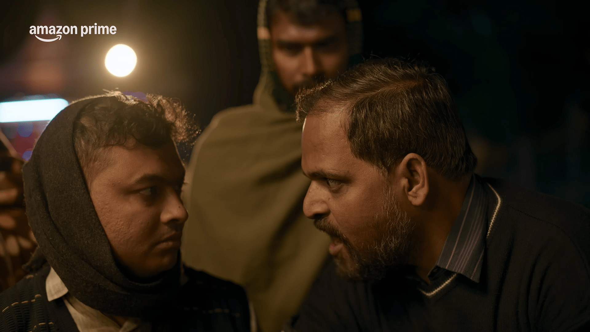 Banarakas in a brown sweater vest looks upward with a concerned expression while sitting outdoors at night, soft yellow light illuminating his face.