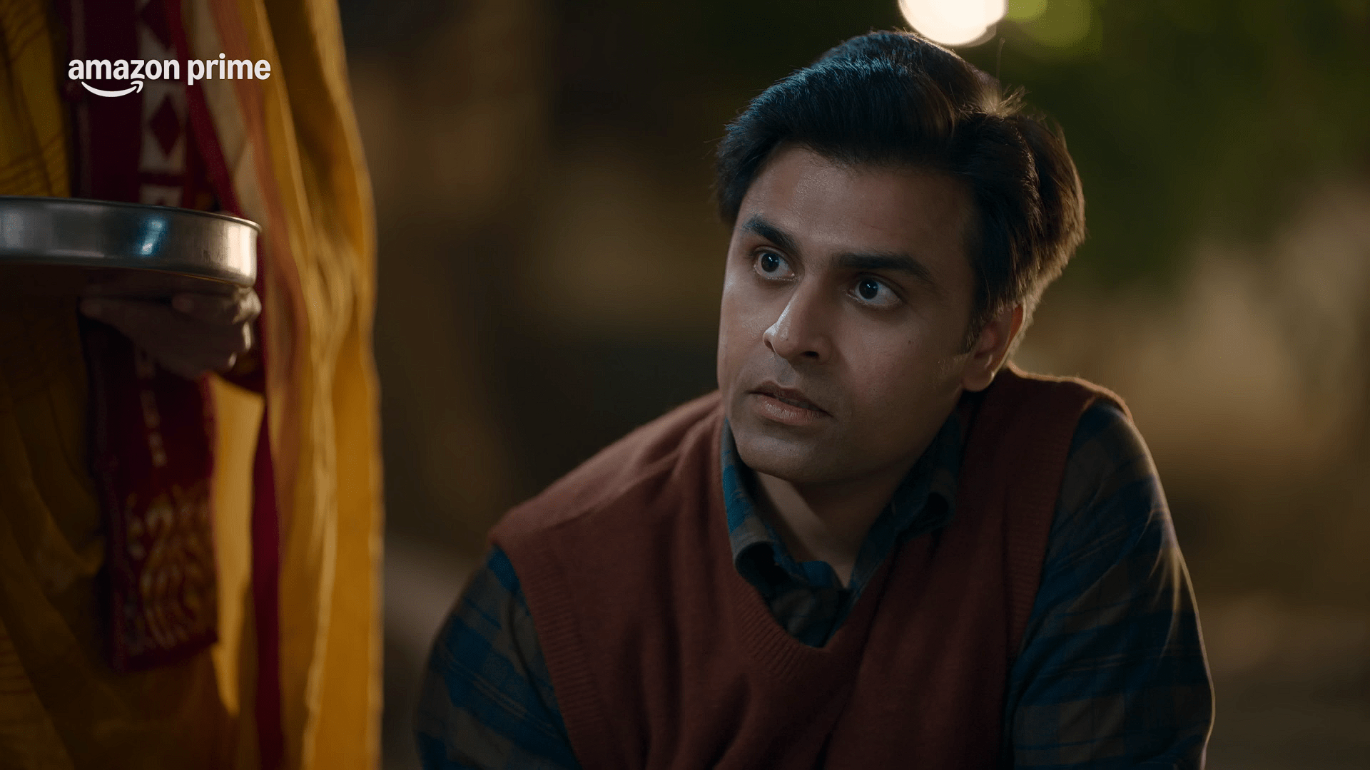 A serious-looking man draped in a brown shawl listens attentively to someone in front of him, standing in a well-maintained garden.