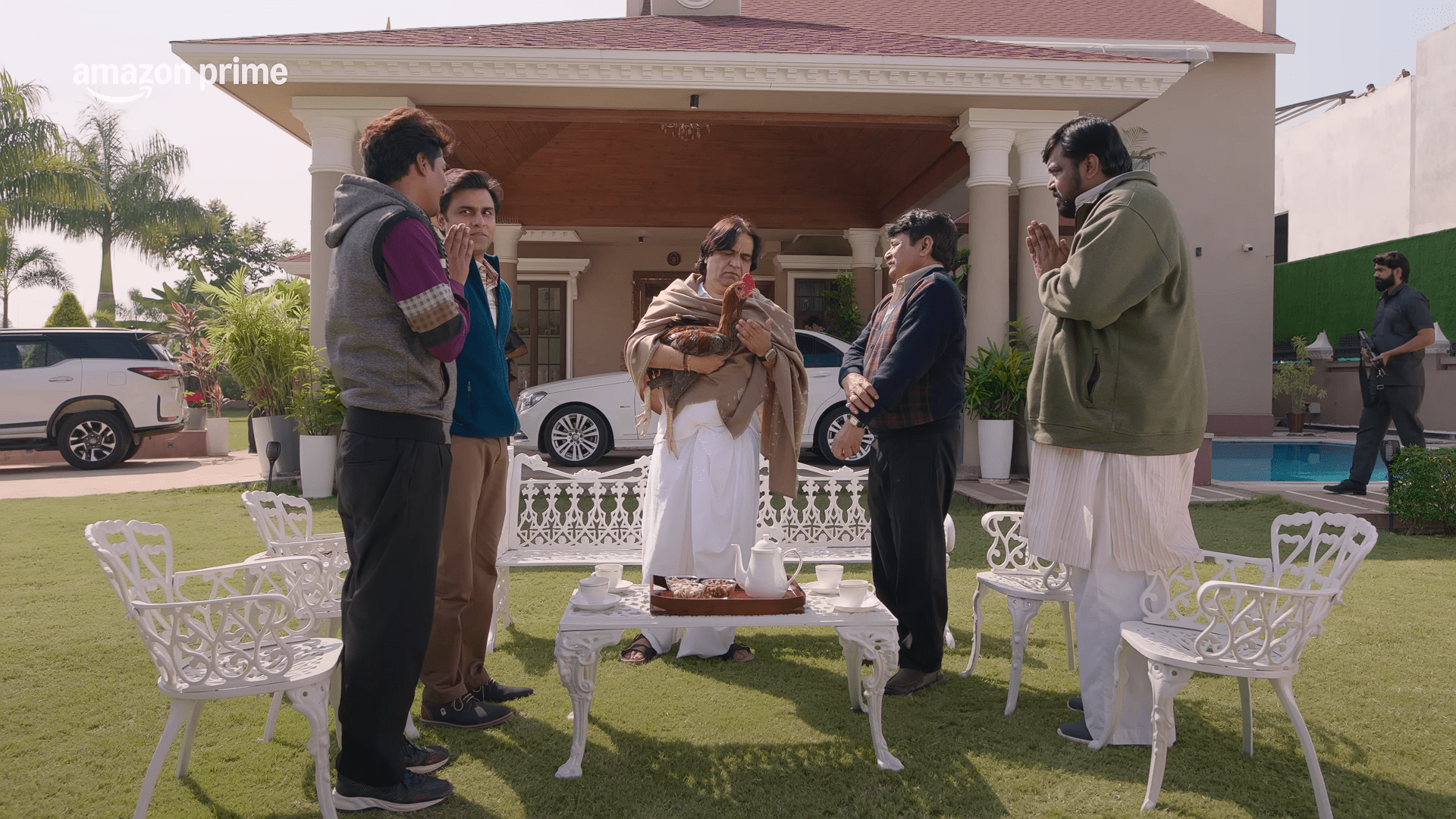 A group of villagers stand respectfully in front of a well-dressed man holding a chicken in his arms, outside a large bungalow with white outdoor furniture and parked cars. (Characters from Panchayat gathered during a formal visit.)