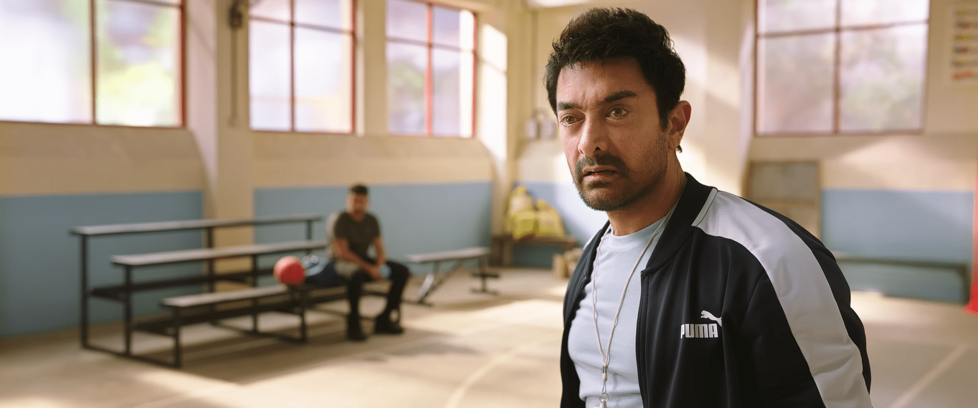 A strict basketball coach looks shocked during practice inside the sports hall, with players sitting on the benches behind him.