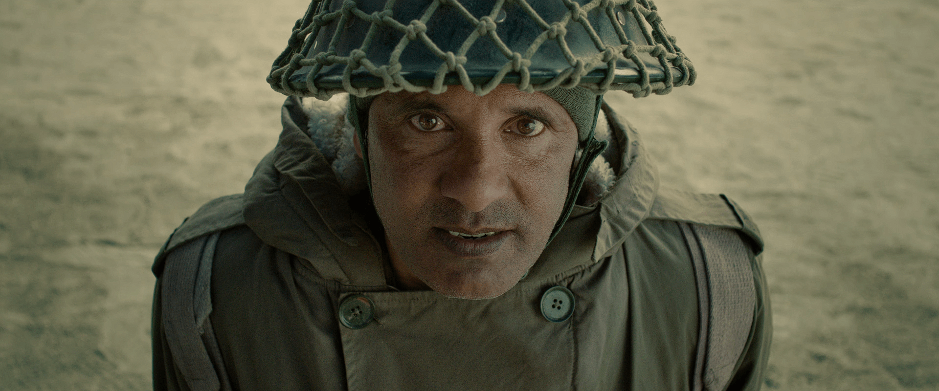 An Indian Soldier wearing a heavy winter coat and army helmet, staring intensely upward with dust-covered skin in a harsh outdoor setting.