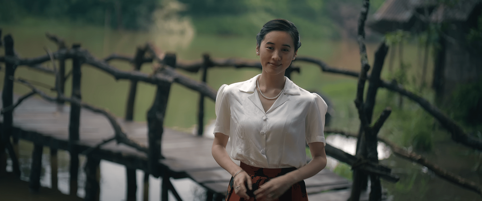 A woman in a white blouse and pearl necklace smiles gently while standing on a rustic wooden bridge overlooking a calm lakeside village setting.