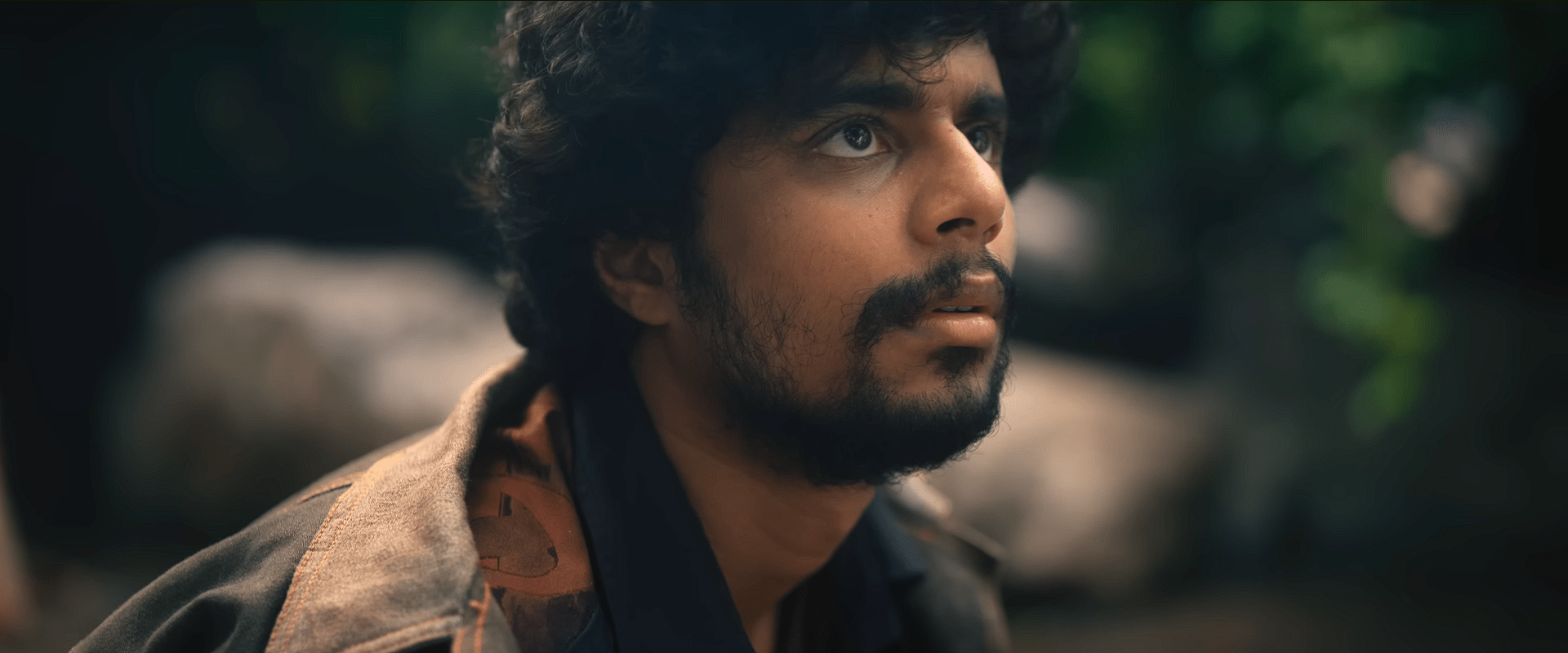 A young man with curly hair and a light beard looks upward with a tense, alert expression, seated outdoors against a blurred forest background.