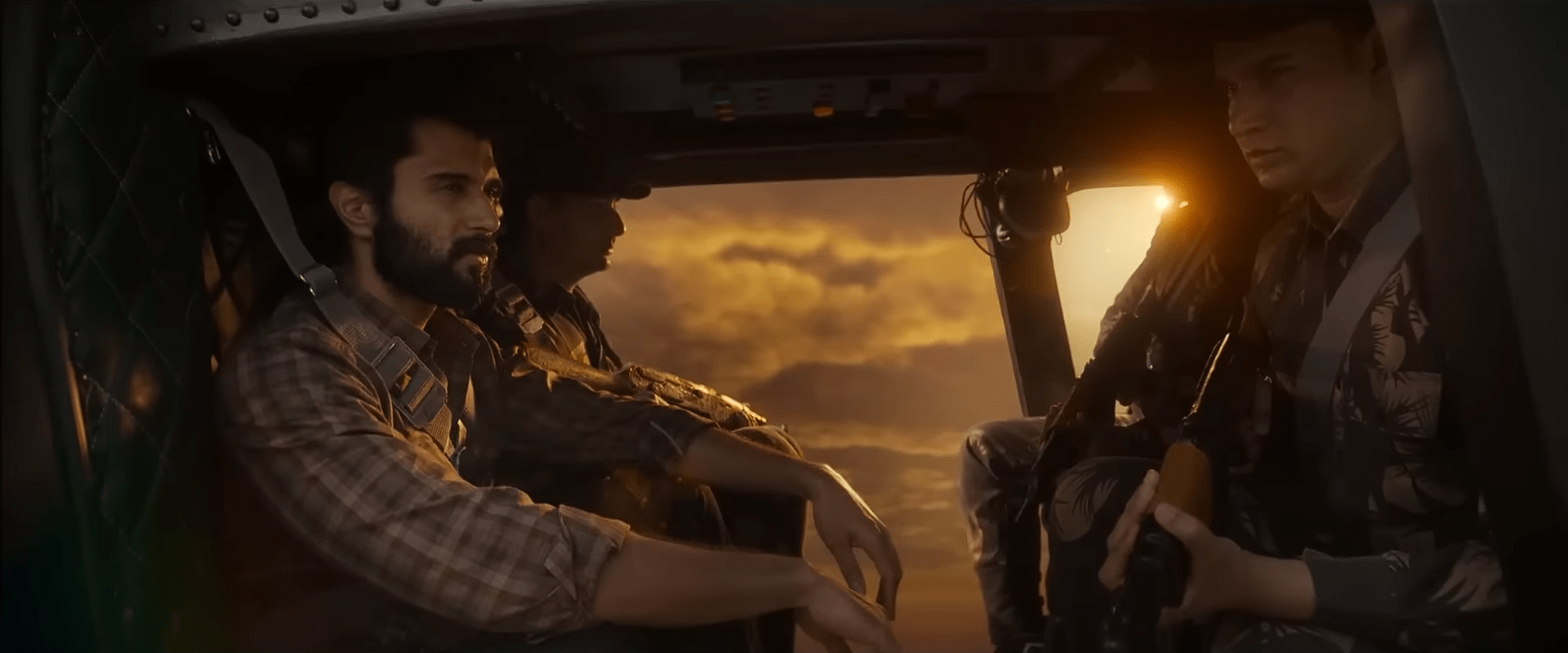 Vijay Deverakonda sits inside a military helicopter with two armed soldiers as the sunset glows through the window.