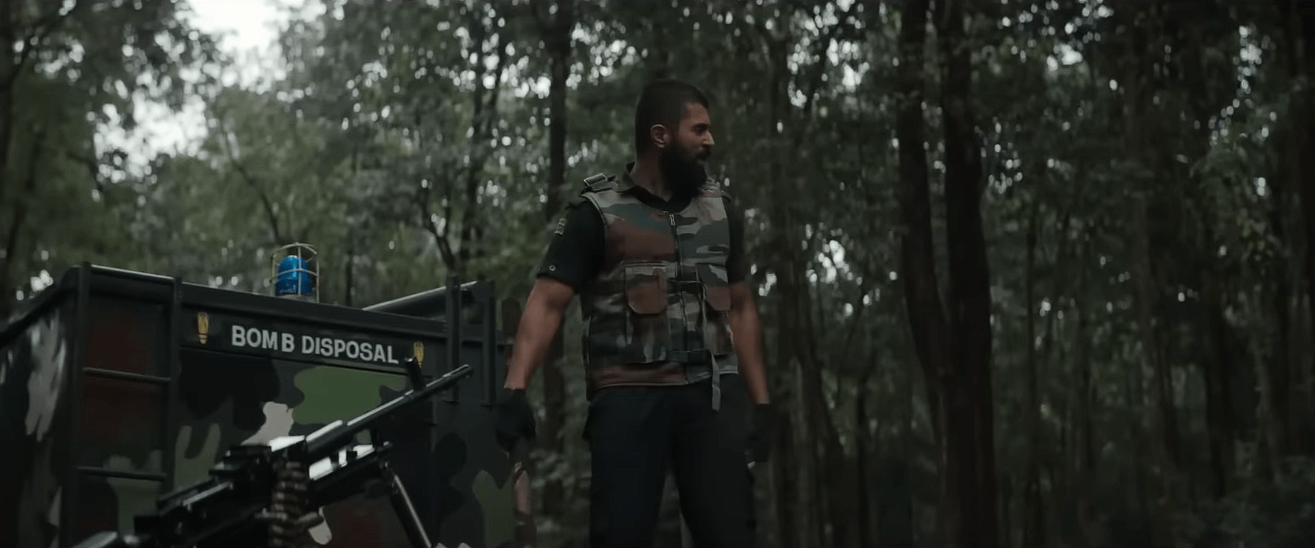 A heavily built soldier in camouflage gear stands beside a bomb-disposal truck in a dense forest.