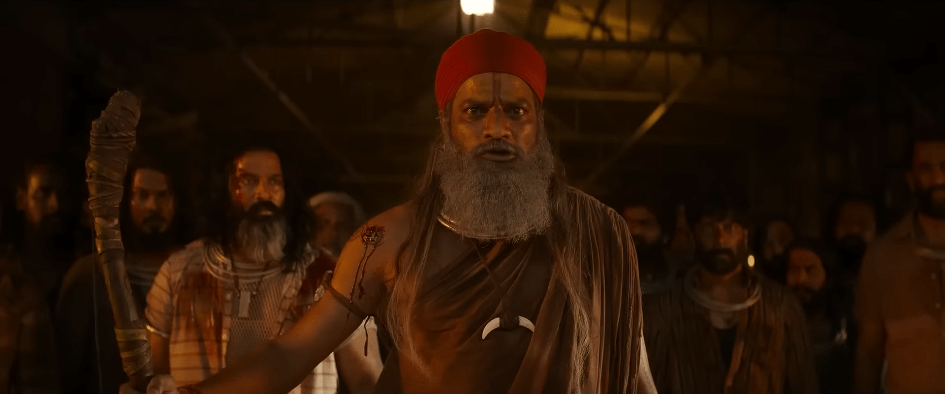 An elderly bearded leader wearing a red headwrap stands at the front of a group of men, holding a wooden staff, in a dark and intense setting.