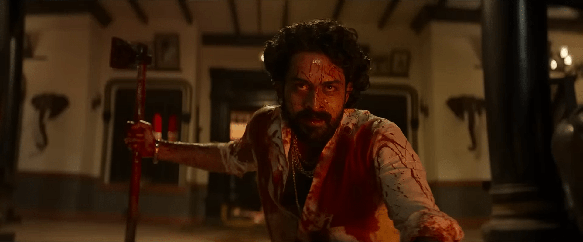 A blood-covered man with curly hair, played by Satyadev, crawls forward on the floor while gripping an axe inside a dimly lit traditional house.