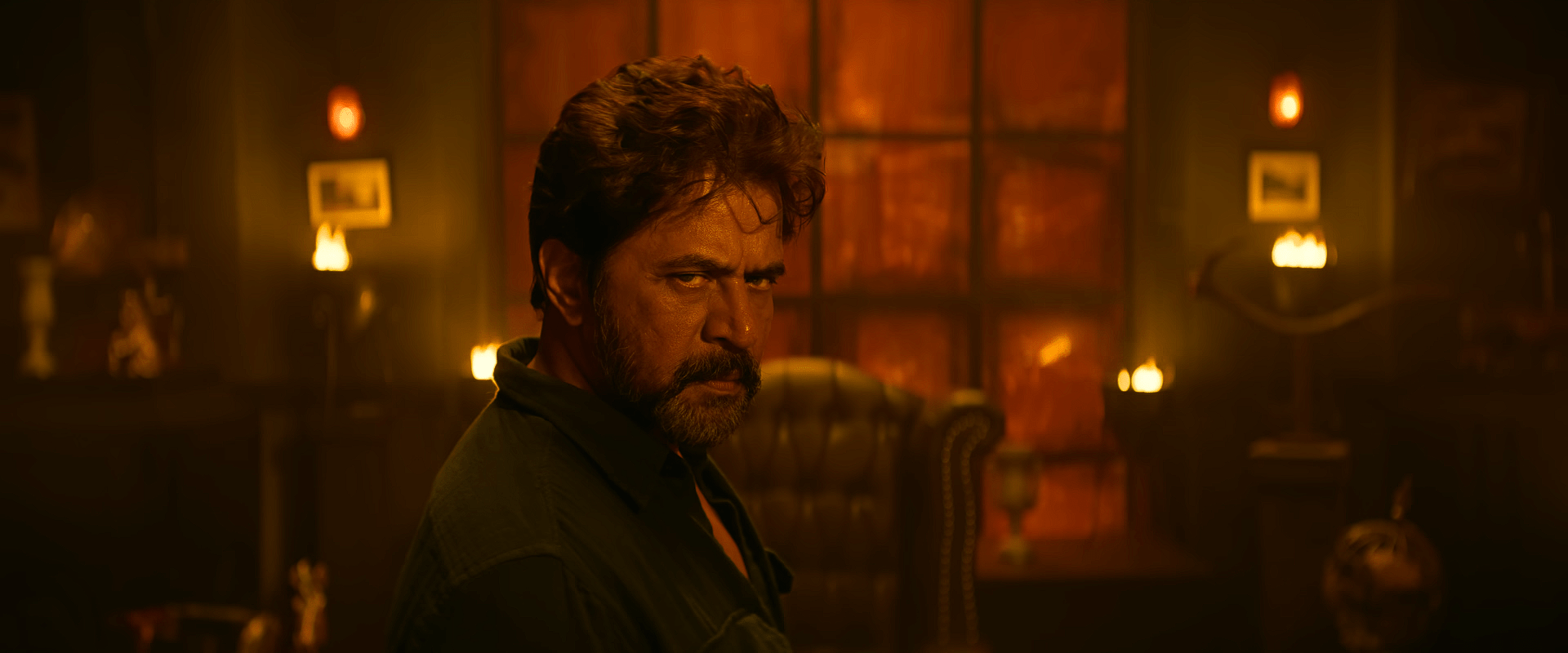 A man stands in a dimly lit room with orange lighting, giving a sharp, intense look over his shoulder, with furniture and lamps in the background.