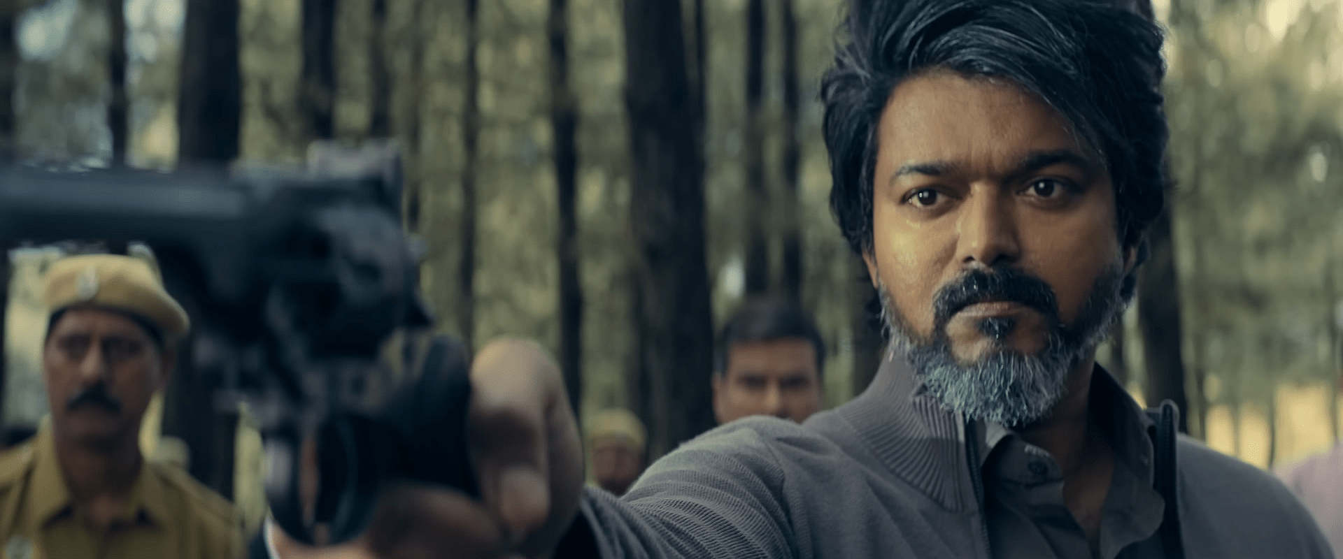 A man with greying hair and beard points a gun forward in a wooded outdoor area, with police officers standing behind him.