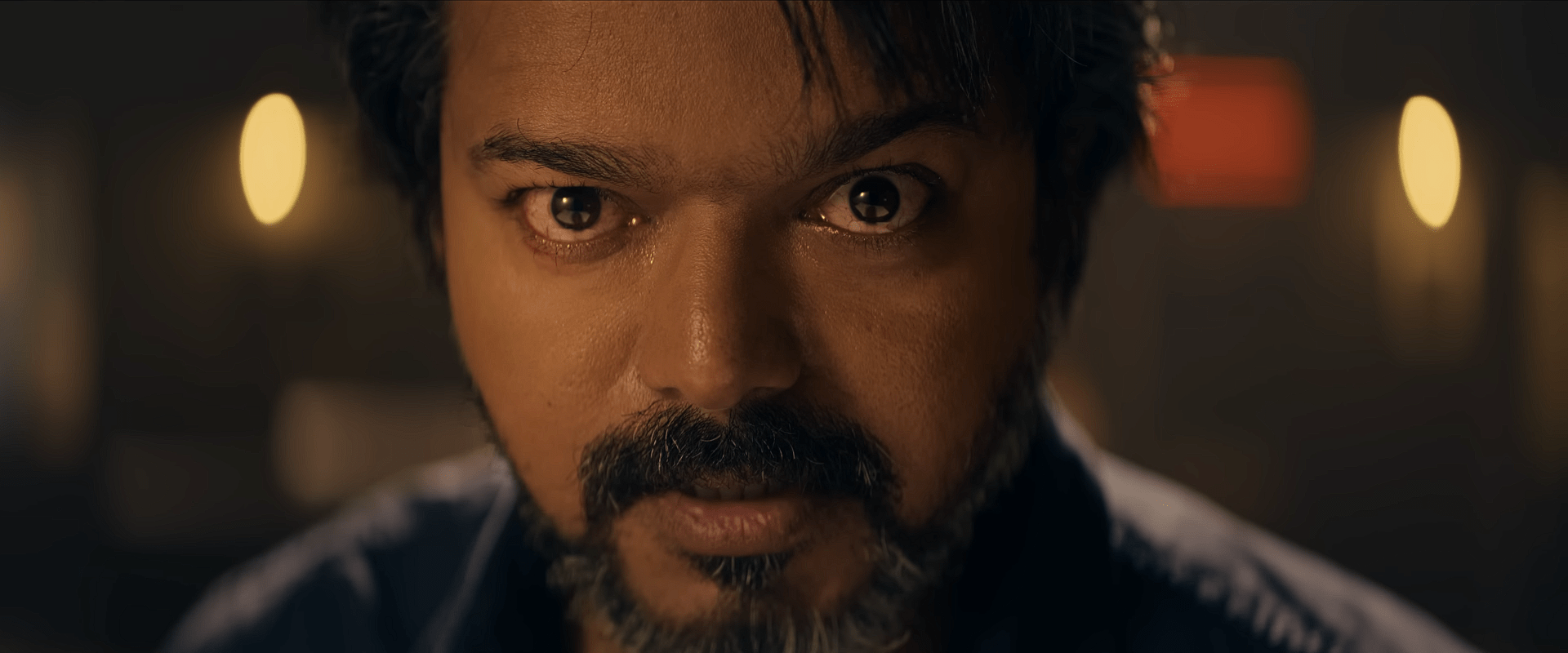 A close-up shot of a bearded man staring intensely into the camera, with warm blurred lights glowing in the background.