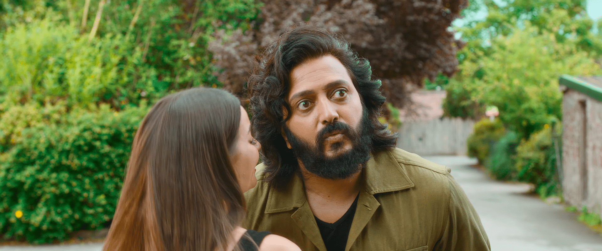 Riteish Deshmukh reacts with a surprised, wide-eyed look as a woman whispers close to his ear in an outdoor lane surrounded by greenery.