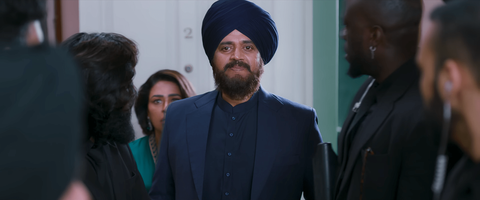 A man in a navy-blue turban and suit stands at a doorway with several people gathered behind him, watching with curiosity.