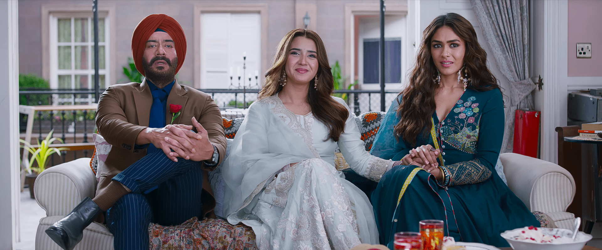 A man in a turban sits beside two women in elegant traditional outfits, all seated on a balcony-side sofa with a spread of food on the table in front of them.