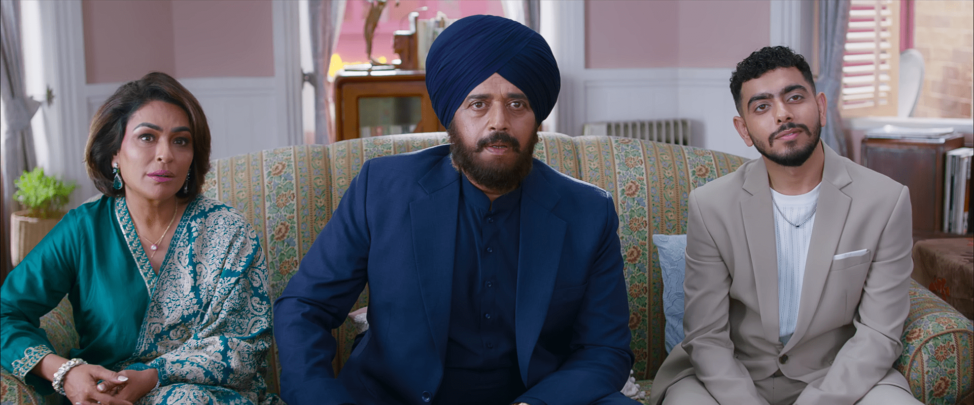 A family of three, dressed in traditional and formal attire, sits on a patterned sofa looking attentively toward the camera.