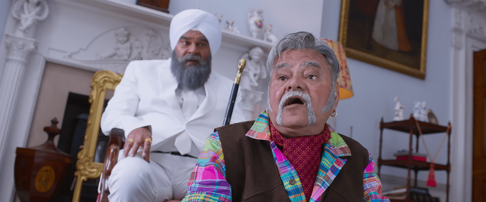An elderly man in a colourful outfit looks shocked while another older man in a white turban and suit watches from behind in an ornate living room.