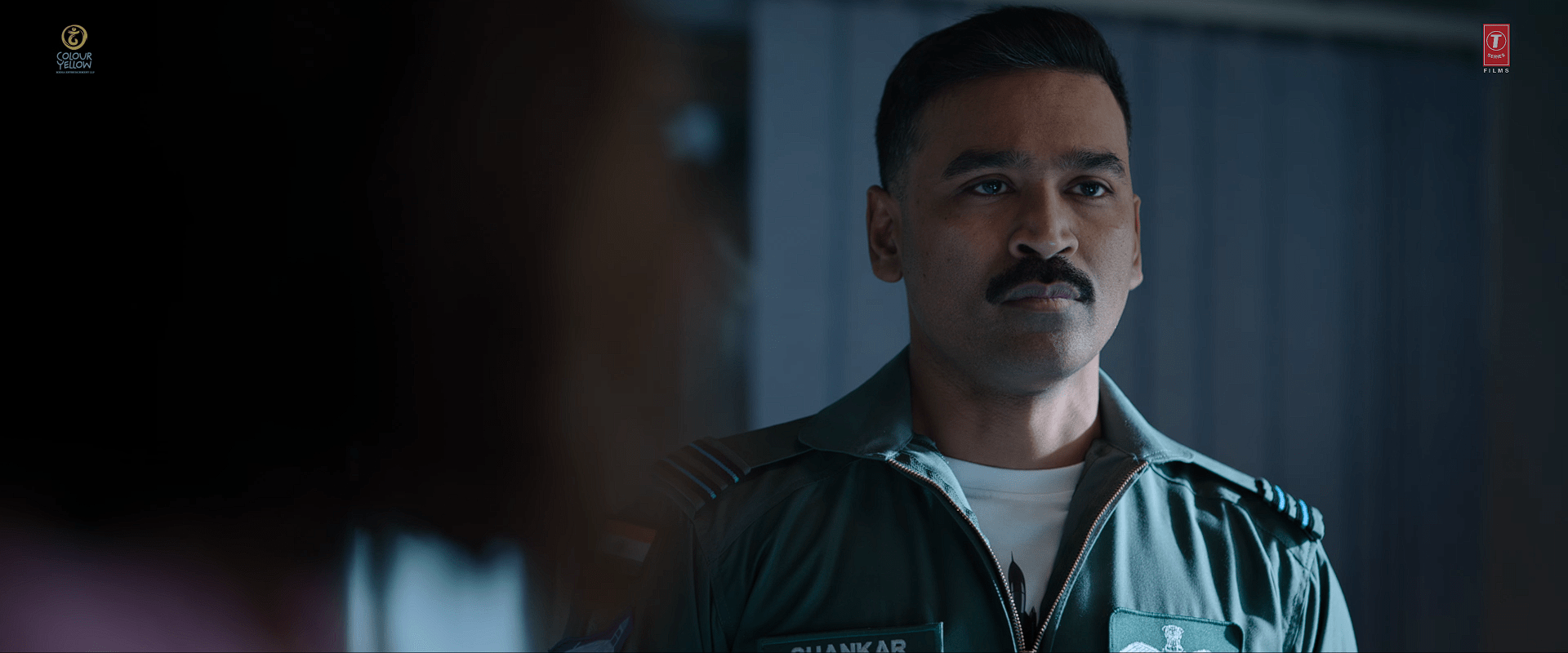 Actor Dhanush in an Air Force uniform with short-cropped hair and a moustache, standing indoors with a serious, composed expression.