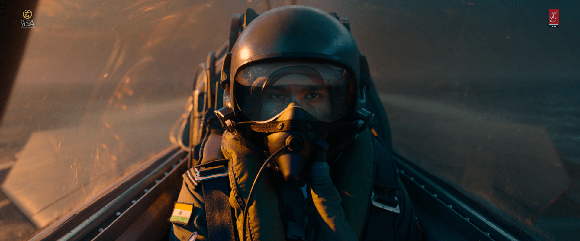 Actor Dhanush wearing a flight helmet and oxygen mask inside a fighter jet cockpit, with glowing light reflecting on the canopy.