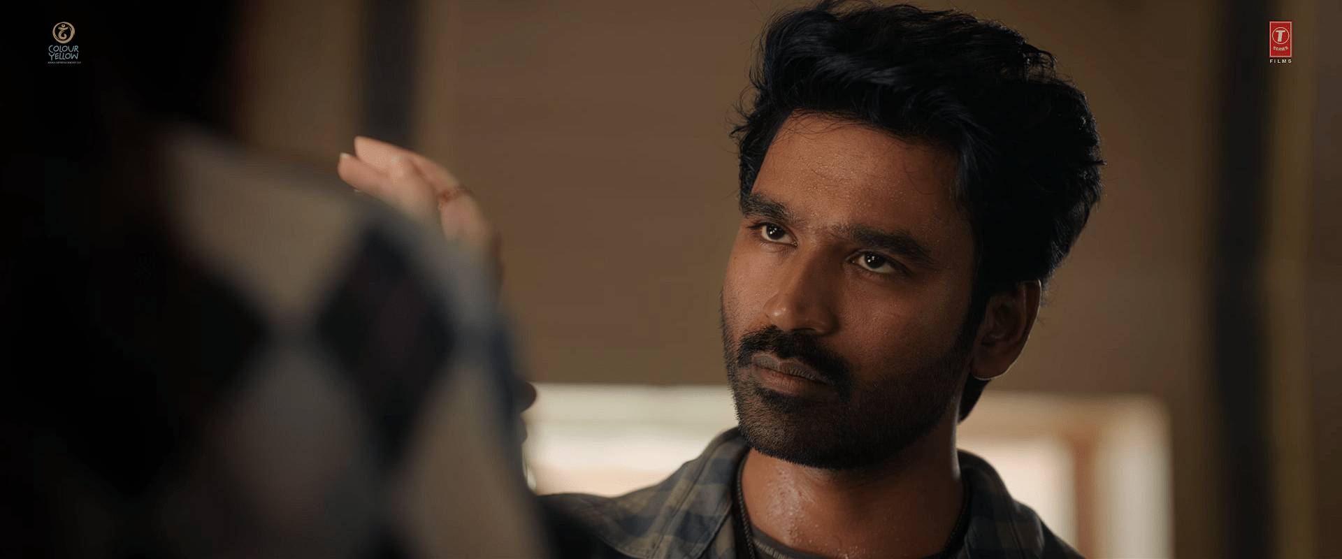A close-up of actor Dhanush gazing upward with a calm yet intense expression as he holds Mukti's hand and looking into her eyes.