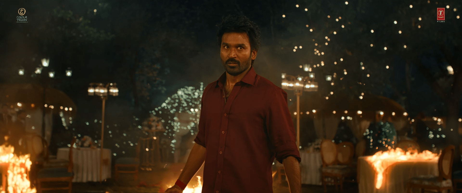 Actor Dhanush in a red shirt standing amid a blazing and chaotic outdoor setting, with fire and sparks surrounding him as he stares intensely ahead.