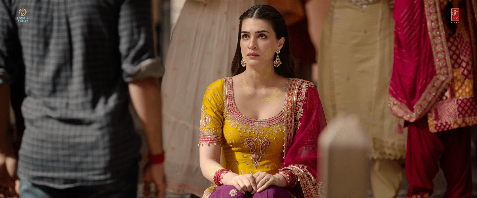 Actress Kriti Sanon dressed in a yellow and pink embellished traditional outfit, sitting during a ritual event and looking upward with a serious expression.