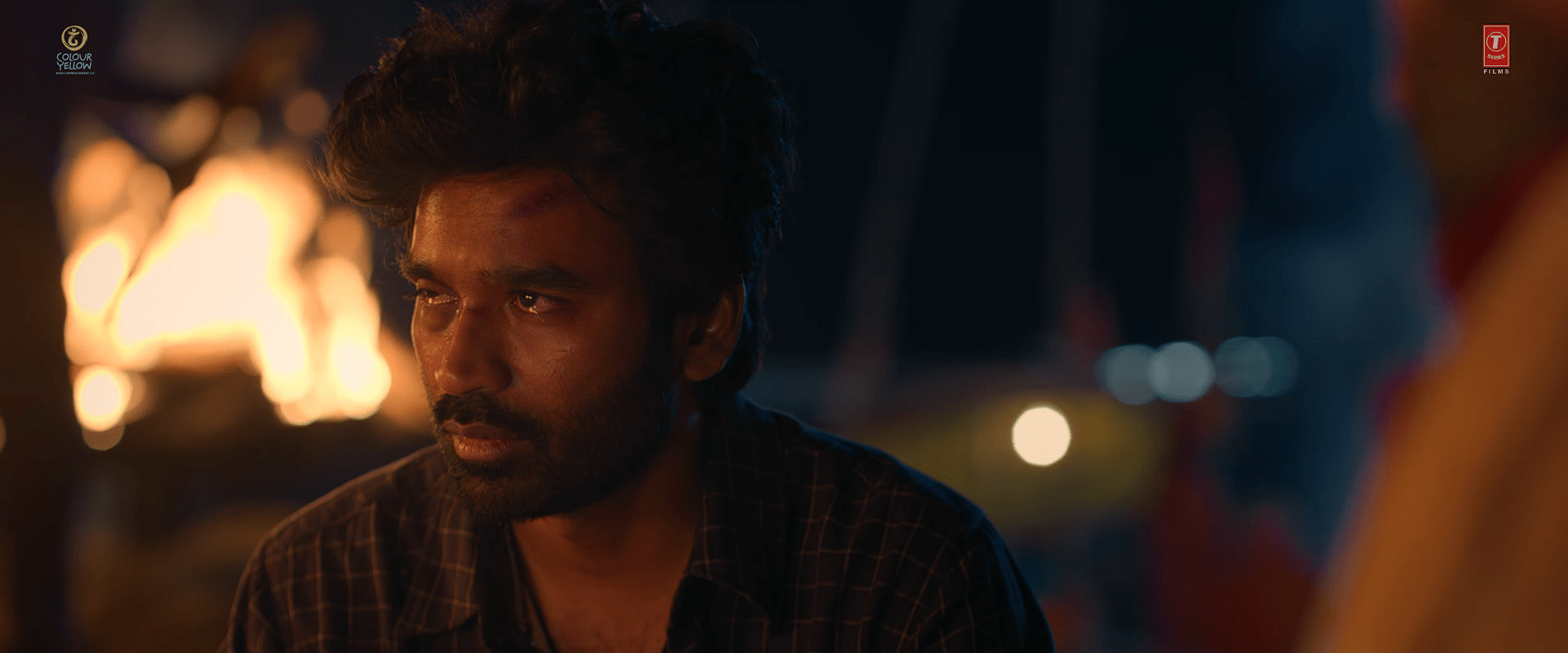 A close-up of actor Dhanush with teary eyes and a bruised forehead, sitting near a burning fire at night, looking distressed.